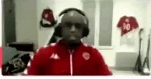 a man wearing headphones and a red jacket is talking on a video call in a dressing room .