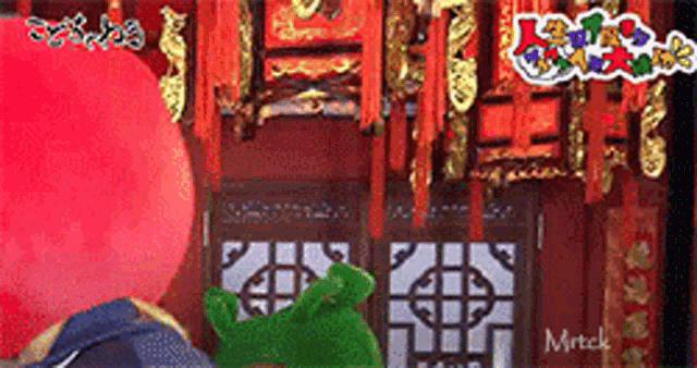 a red balloon and a green frog are standing in front of a chinese building .