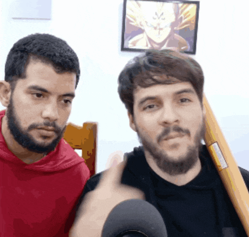 a man with a beard is sitting next to another man with a picture of dragon ball z on the wall behind him