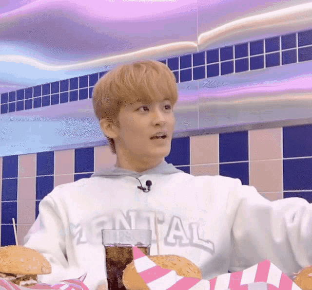 a young man wearing a mental sweatshirt sits at a table with burgers and a glass of soda