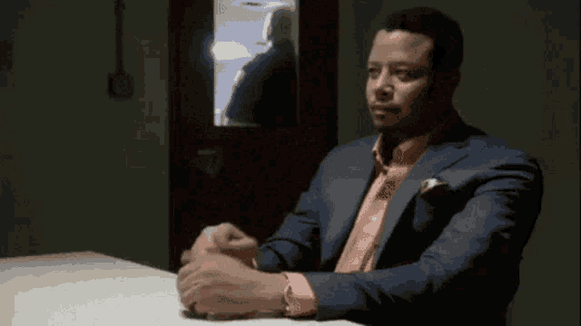 a man in a suit and pink shirt is sitting at a table in front of a window .