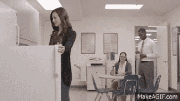 a woman is opening a refrigerator in an office while a man stands behind her .