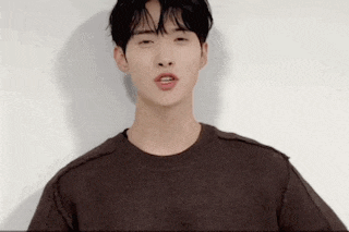 a young man wearing a brown sweater is standing in front of a white wall and looking at the camera .