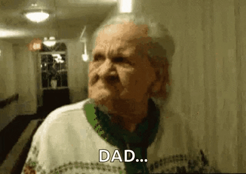 an elderly woman is standing in a hallway and talking to someone while wearing a sweater and scarf .