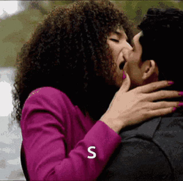 a woman in a pink jacket kisses a man in a black jacket with the letter s above them
