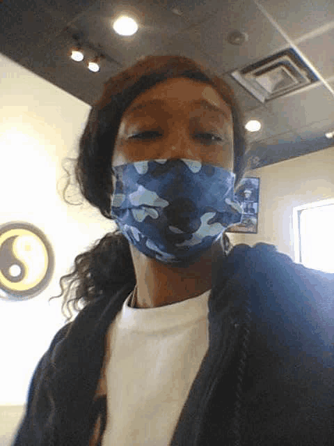 a woman wearing a blue camo face mask stands in front of a wall with a yin yang symbol on it