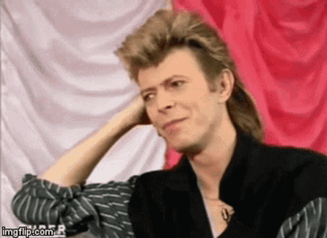 a man with a mullet and a striped shirt is sitting in front of a red curtain .