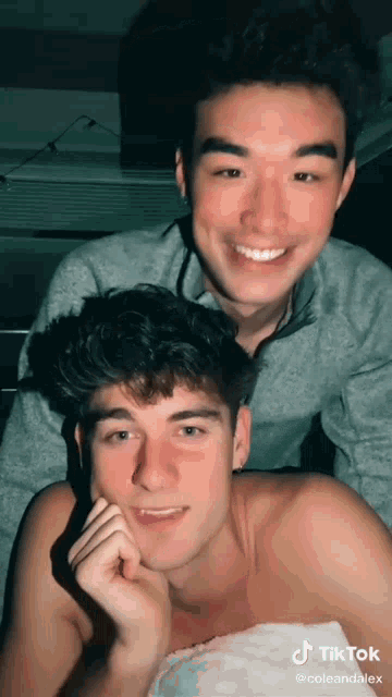 two young men are posing for a picture and one of them is smiling for the camera while the other is wrapped in a towel .