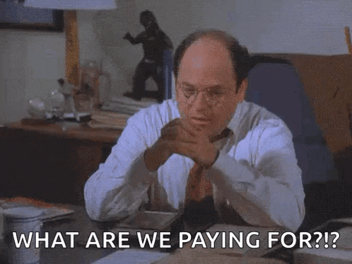 a bald man in a white shirt and tie is sitting at a desk and asking what are we paying for .