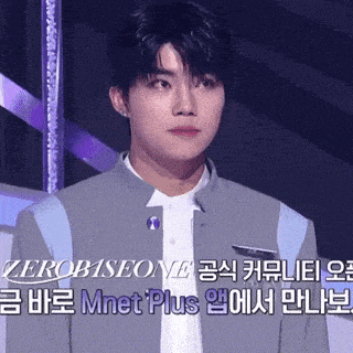 a young man in a gray jacket and white shirt stands in front of a sign that says mnet plus