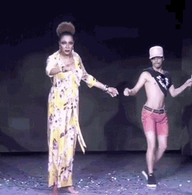 a woman in a yellow dress is holding hands with a shirtless man in pink shorts