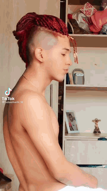 a shirtless man with red braids and a shaved head is sitting in front of a shelf .