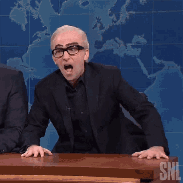 a man in a suit and glasses is sitting at a table with the word snl on it