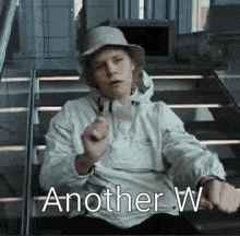 a man wearing a hat and a white jacket is sitting on a set of stairs and giving the middle finger .