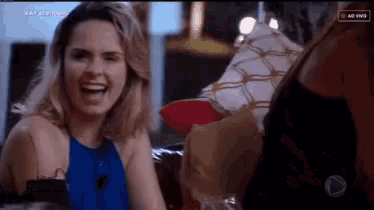 a woman in a blue top is laughing while sitting on a couch next to another woman .
