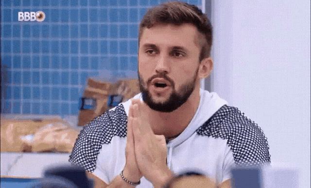 a man with a beard is sitting at a table with his hands folded in front of him .