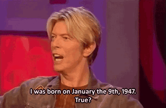 a man is sitting in front of a red curtain and talking about his birth date .