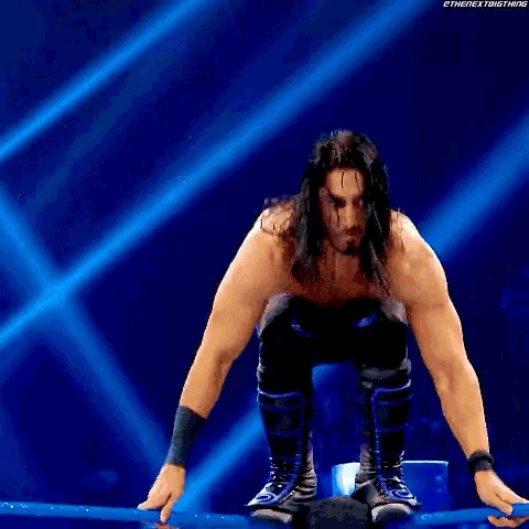 a wrestler with long hair is kneeling down on a blue rope .