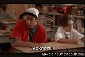 a man in a red shirt is sitting at a desk in a classroom and says jackpot !!!