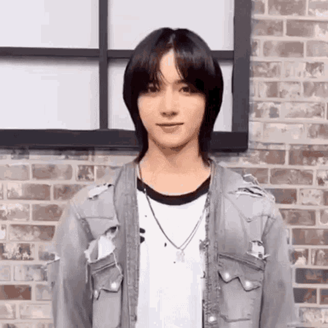 a young man with long black hair is standing in front of a brick wall wearing a denim jacket and a white shirt .