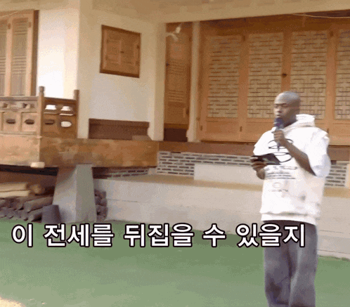 a man stands in front of a house holding a microphone with korean writing on the bottom