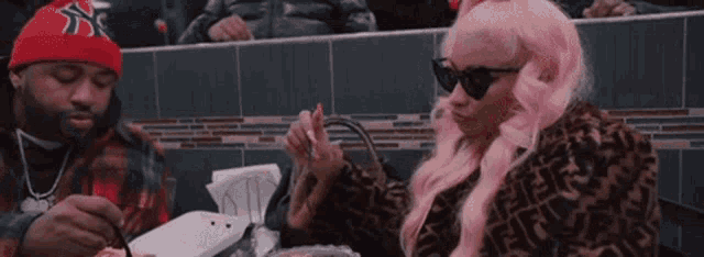 a woman with pink hair and sunglasses is sitting at a table eating food with a fork .