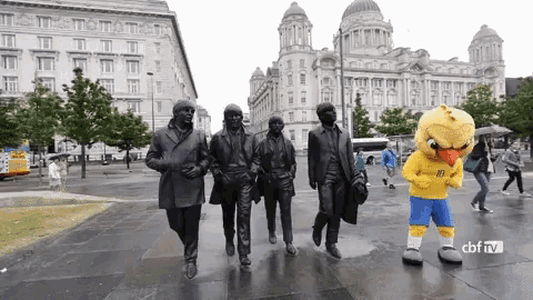 a statue of the beatles walking down a street with a mascot in front of them