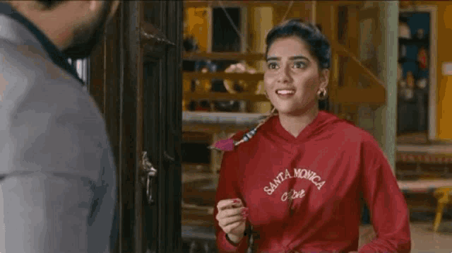 a woman in a red santa monica hoodie is holding a rose and talking to a man in a suit .