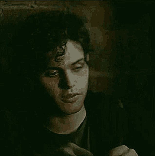 a man with curly hair is sitting in front of a brick wall and smoking a cigarette .