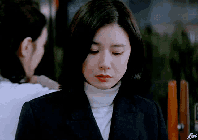 a woman in a black jacket and white turtleneck looks down at something