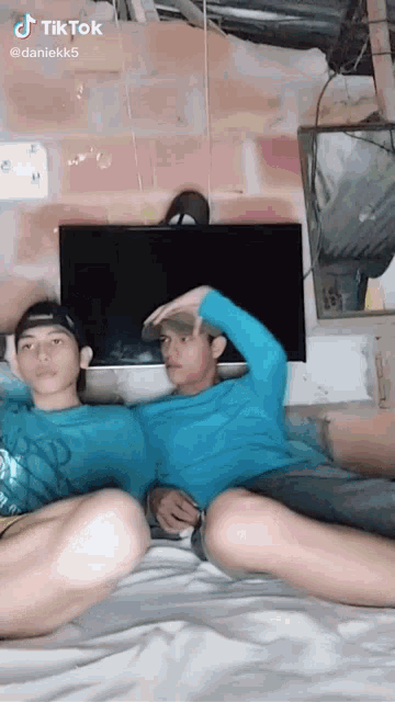 two young men are sitting on a bed with their legs crossed and a television in the background .