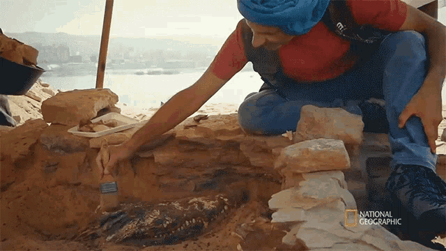 a man in a red shirt is digging in the sand with a national geographic logo above him