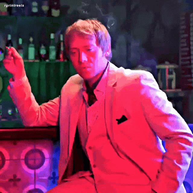 a man in a suit is smoking a cigarette in front of a bar with bottles behind him