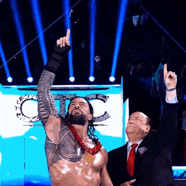 a man in a suit and tie stands next to a wrestler who is pointing up