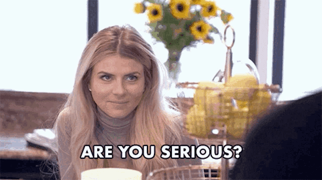 a woman is sitting at a table with a vase of sunflowers in the background and says are you serious .