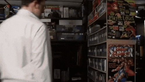 a man in a white shirt is standing in front of a shelf full of video games including super smash bros .