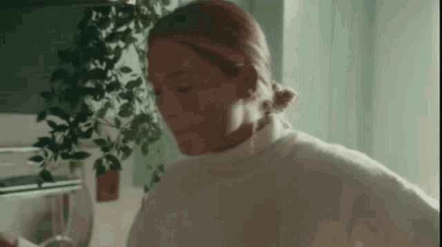 a woman in a white sweater is standing in front of a plant in a kitchen .