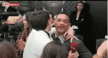 a man in a suit is being interviewed by a woman while another man kisses him on the cheek .