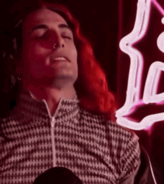 a man with red hair and headphones is sitting in front of a neon sign with his eyes closed .