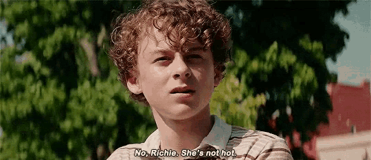 a young man with curly hair is standing in front of a tree and talking to someone .