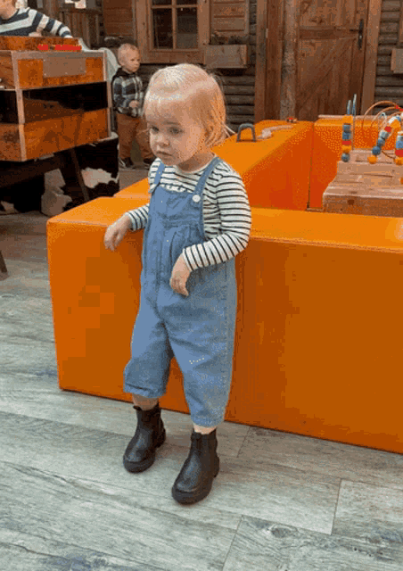 a little girl wearing overalls and black boots stands on an orange cube