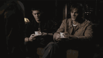 two men sit on a couch holding cups of coffee in front of a lamp