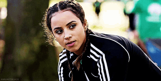 a woman with braids and a nose ring is wearing a black adidas track jacket .