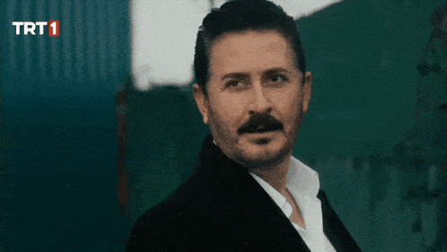 a man with a beard is looking at the camera with a trt1 logo behind him