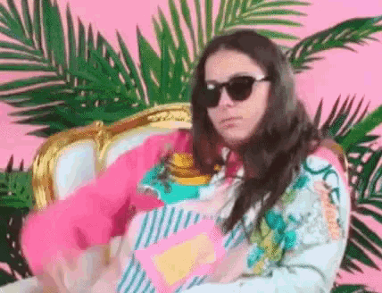 a woman wearing sunglasses is sitting in a chair with a pink flamingo in her lap .