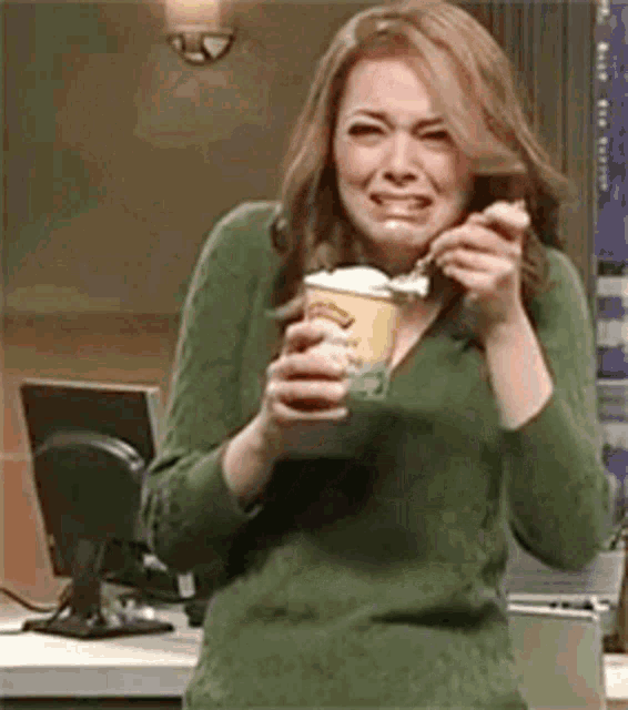 a woman in a green sweater is holding a cup of ice cream and eating it .