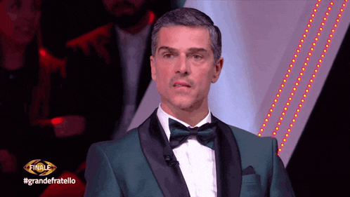 a man in a green tuxedo and bow tie is standing in front of a sign that says finale grandefratello