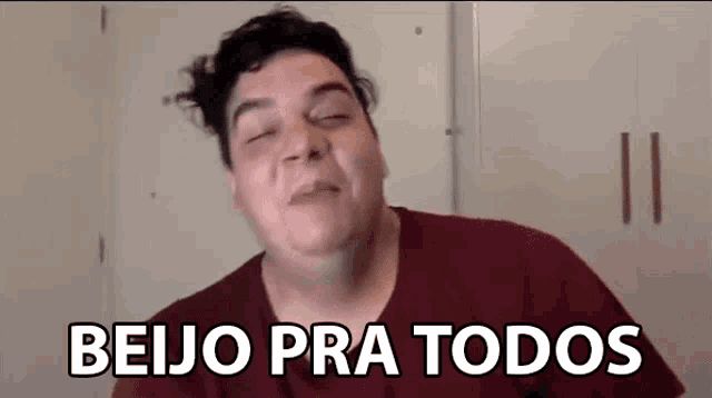 a man in a red shirt is making a funny face and the words beijo pra todos are behind him .