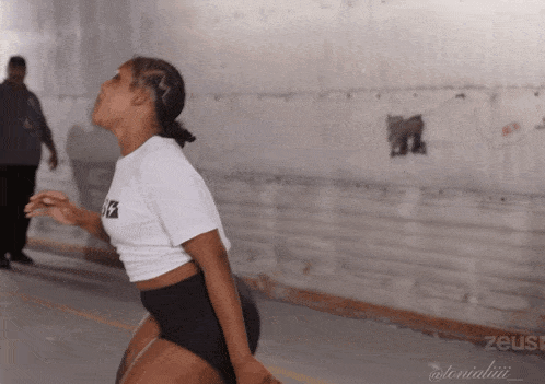 a woman in a white t-shirt and black shorts is dancing in front of a wall that says zeus