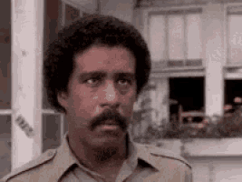 a man with a mustache and curly hair is standing in front of a building and looking at the camera .